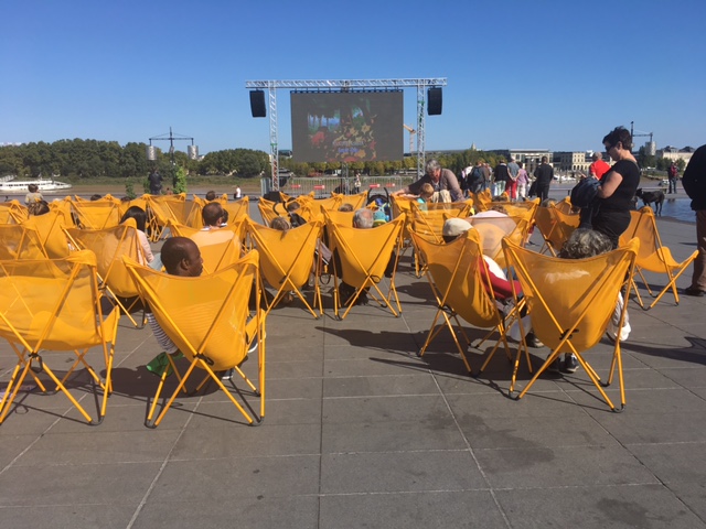 Kids and all ages enjoy a movie in the broad daylight on a warm afternoon on the river bank by the reflecting pool with sprinklers that shoot from the tiled floor.